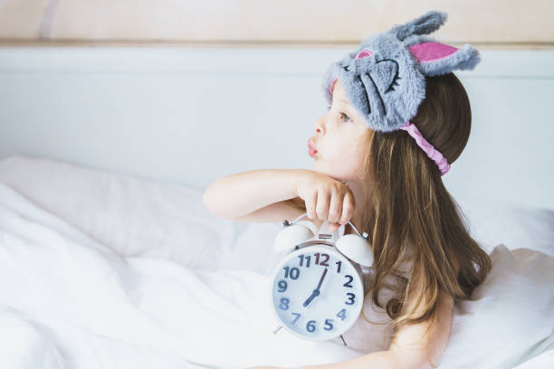 Little girl wakes up in morning under white blanket wearing grey bunny plush sleep mask with alarm clock in her hands. Child sleep regime hours. Early rise to kindergarten. Mom's daily rest routine