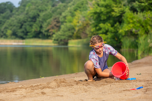 Close-up of a ten year old girl pouring water from a red bucket into the sand at the beach.