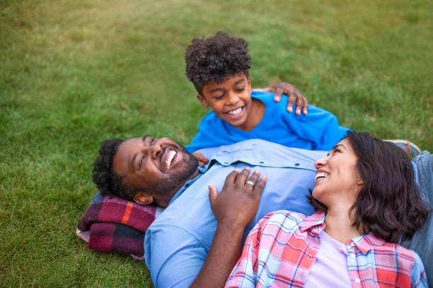 multicultural mixed race multi-ethnic family with one child laying on grass laughing - offspring child lying on back parent imagens e fotografias de stock