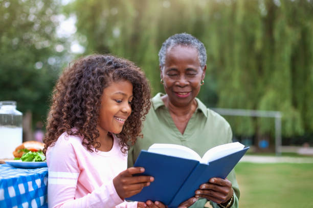 granddaughter reading book with grandmother at public park picnic - grandparent reading grandmother child imagens e fotografias de stock