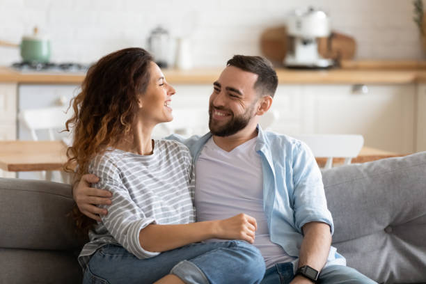 Happy young couple of man and woman embracing. Happy young couple of man and woman embracing look at each other sitting on couch in living room. Attractive smiling husband and wife laughing having fun free time together. two parents stock pictures, royalty-free photos & images
