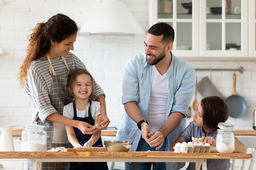 Caring father and mother playfully teaching little children to cooking pastry standing at table in kitchen. Happy smiling dad and mom with son and daughter playing with flour preparing dinner.