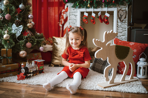 Smiling little girl is sitting next to the fireplace , dressed with red dress    sitting on the carpet , Christmas holidays and Christmas decoration