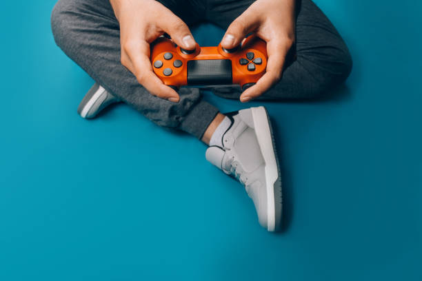 Young guy plays video games A young guy plays video games in his hands holding a red gamepad on a blue background, sitting in gray sneakers computer game control stock pictures, royalty-free photos & images