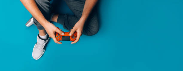 Young guy plays video games A young guy plays video games in his hands holding a red gamepad on a blue background, sitting in gray sneakers, long banner game online web rpg stock pictures, royalty-free photos & images