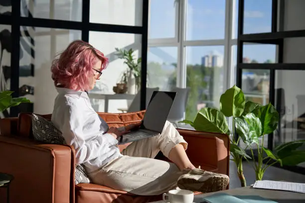 Stylish creative hipster young woman, teen girl with pink hair using pc laptop computer browsing internet work learning online sitting on sofa in modern home office lit with morning sunshine.