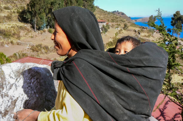 Taquile, Peru - 07/24/2006: Smiling mother carrying her baby on her back in a traditional Peruvian carrying cloth called manta or awayo.