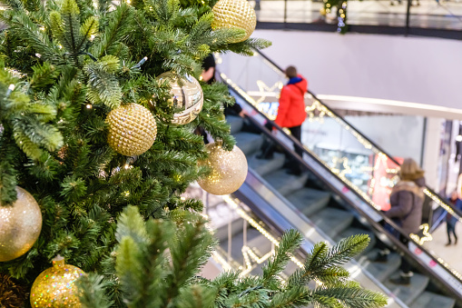 Golden Christmas balls and garland on a decorated fir tree in Shopping Mall. Blurred people on escalator, in festive business center make New Year's purchases,.