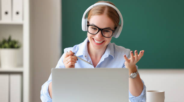 Female teacher during online lesson in classroom Happy woman in headphones and glasses smiling and talking with students while making video call in classroom online tutors stock pictures, royalty-free photos & images