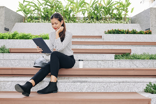 Positive young multi-ethnic businesswoman sitting on steps outdoors during break, drinking coffee and reading novel on digital tablet