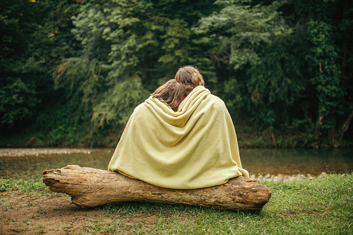 Brother and sister camping in the woods on the mountain. They sitting in front of their blue tent on a tree trunk, hugging and covered with a yellow blanket