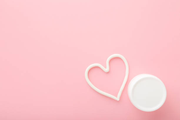istock Heart shape created from cream. White jar on light pink table background. Pastel color. Care about face, hands, legs and body skin. Closeup. Empty place for text or logo. Top down view. 1269809426