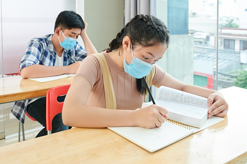 Vietnamese school children in medical masks reading books and writing in copybooks during lesson