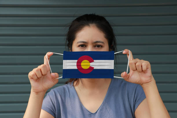 A woman and hygienic mask with Colorado flag pattern in her hand and raises it to cover her face on dark green background. stock photo