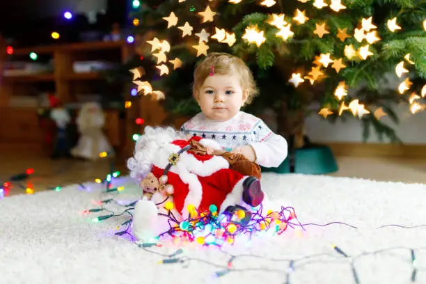 Adorable baby girl holding colorful lights garland in cute hands. Little child in festive clothes decorating Christmas tree with family. First celebration of traditional holiday called Weihnachten.