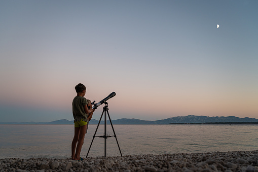 Two brothers standing on beautiful pebble beach by the sea looking to the stars with a telescope.