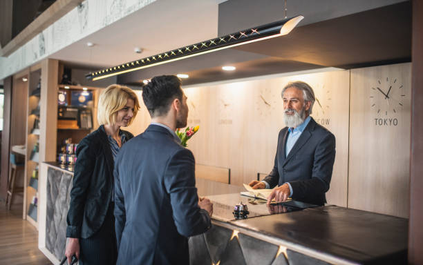 Hotel Receptionist Conversing with Guests Checking In Mature hotel receptionist with full beard smiling as he talks with young male and female business travelers checking in for overnight stay. guest book photos stock pictures, royalty-free photos & images