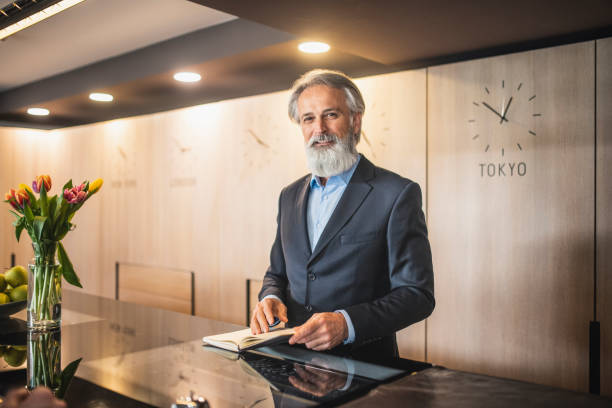 Bearded Hotel Receptionist Helping Guest at Front Desk Candid portrait of Caucasian man in mid 50s wearing suit with open collar shirt and smiling at camera while assisting guest in modern hotel. guest book photos stock pictures, royalty-free photos & images