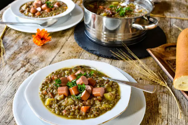 fresh and homemade hearty lentil soup / stew with sausages served on a white soup plate and cooking pot on a wooden table at home - ready to eat