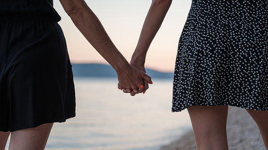 Closeup view of lesbian gay couple holding hands walking on pebble beach in the evening.
