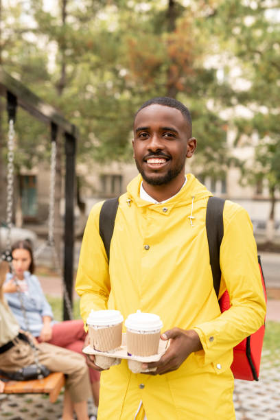 istock Happy young contemporary male courier in yellow jacket holding two drinks 1269814503