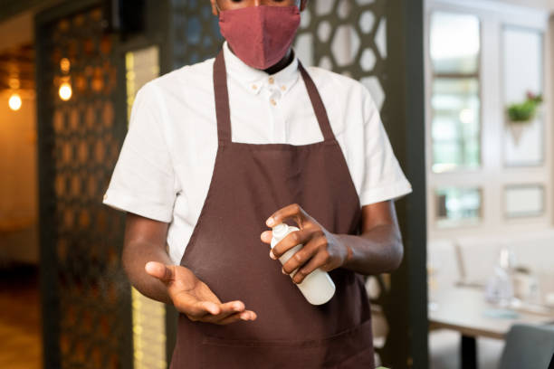 istock Young waiter in uniform and protective mask spraying santitizer on hands 1269814541