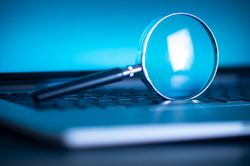 Laptop computer with magnifying glass as a symbol for searching information on the internet. blue tinted image with selective focus on the glass of a magnifying glass that is upright on the laptop