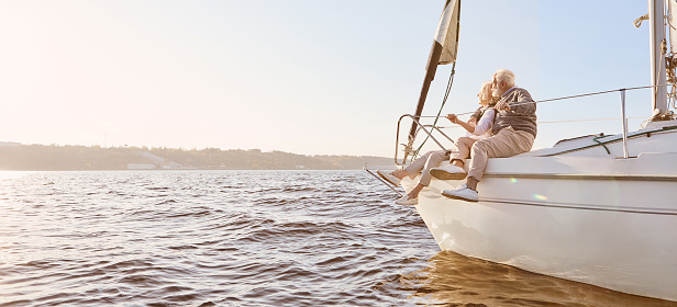 A happy senior couple sitting on the side of a sail boat on a calm blue sea. Man hugging his woman while enjoying view. Sailing, age, tourism, travel and people concept. Web Banner