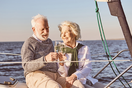 Close up of happy senior couple sitting on the side of sail boat or yacht deck floating in sea. Man and woman drinking wine, enjoying the view. Sailing, travel and people concept. Horizontal shot