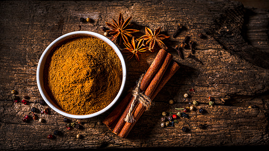 Scented spices background: top view of a white bowl filled with cinnamon powder shot on rustic wooden table. Cinnamon sticks, star anise, cloves and peppercorns are all around the bowl.  Predominant color is brown. The composition is at the left of an horizontal frame leaving useful copy space for text and/or logo at the right. High resolution 16:9 format studio digital capture taken with SONY A7rII and Zeiss Batis 40mm F2.0 CF lens