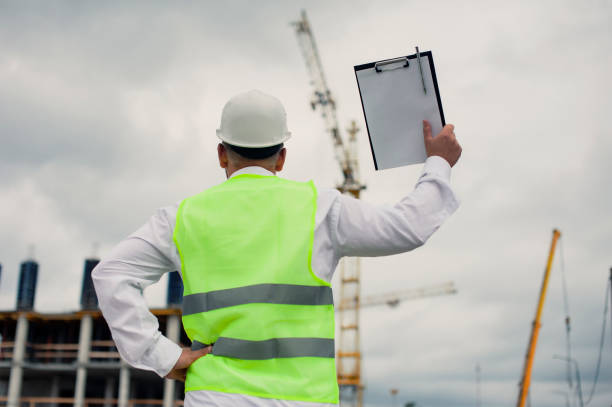 istock Chief in a vest and a construction helmet with documents in his hands, against the background of a tower crane and the sky with clouds. House construction inspection. 1269840271