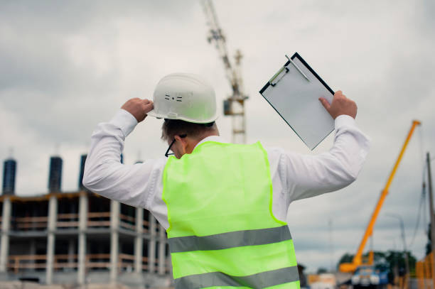 istock Chief in a vest and a construction helmet with documents in his hands, against the background of a tower crane and the sky with clouds. House construction inspection. 1269840280