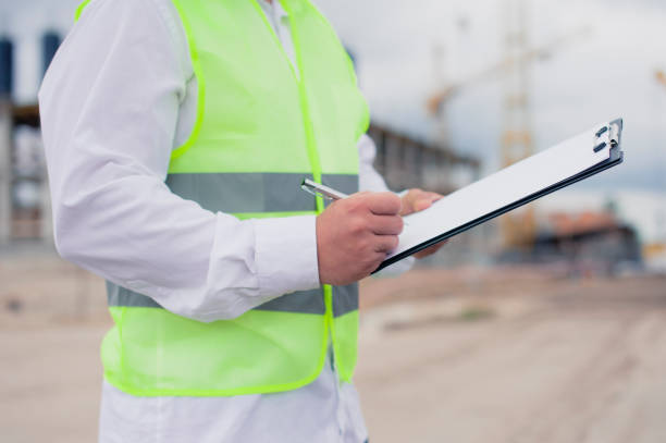 istock Chief in a vest and a construction helmet with documents in his hands, against the background of a tower crane and the sky with clouds. House construction inspection. 1269840322