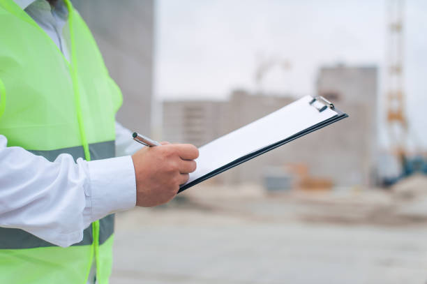 istock Chief in a vest and a construction helmet with documents in his hands, against the background of a tower crane and the sky with clouds. House construction inspection. 1269841325