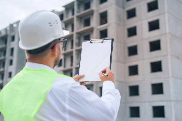 istock Chief in a vest and a construction helmet with documents in his hands, against the background of a tower crane and the sky with clouds. House construction inspection. 1269841331
