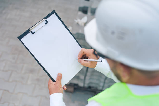 istock Chief in a vest and a construction helmet with documents in his hands, against the background of a tower crane and the sky with clouds. House construction inspection. 1269841336