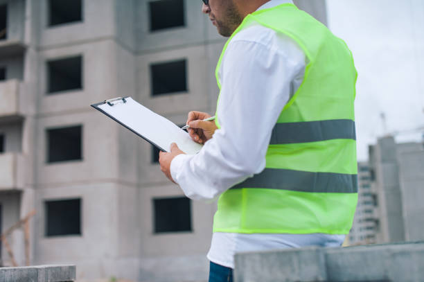 istock Chief in a vest and a construction helmet with documents in his hands, against the background of a tower crane and the sky with clouds. House construction inspection. 1269841338