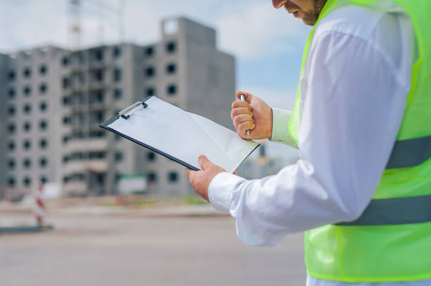 istock Chief in a vest and a construction helmet with documents in his hands, against the background of a tower crane and the sky with clouds. House construction inspection. 1269841352