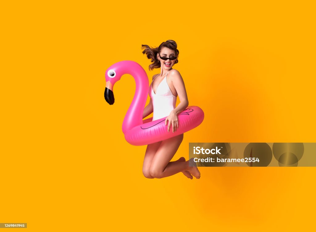 Happy young woman jumping on yellow background dressed in swimwear holding flamingo rubber ring beach. Summer Stock Photo
