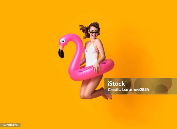 Happy Young Woman Jumping On Yellow Background Dressed In Swimwear Holding Flamingo Rubber Ring Beach Stock Photo - Download Image Now