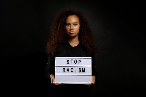 Portrait of young beautiful black woman standing in the dark with anti racist slogan. Protesting female holding a light box, black wall background. Close up, copy space.