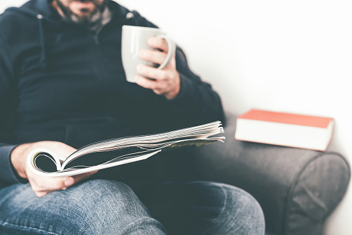 close-up of caucasian man reading magazine or journal on sofa while holding a cup of coffee