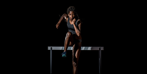 Front view of an African-American female athlete running away from a hurdle towards the camera.