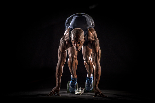 Front view of a muscular African-American sprinter at the starting line ready to start with his feet on the starting blocks and looking in front of him. Black background.