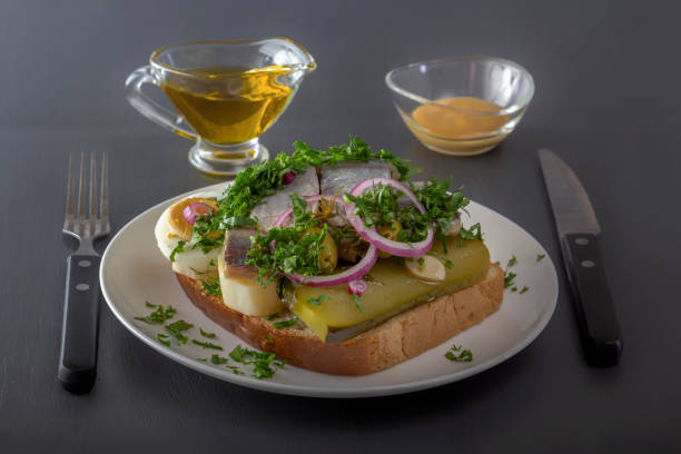 istock Sandwich with herring fillets, onions, pickled cucumbers, eggs and herbs on a white plate on a dark table against the background of a sauce bowl and butter dish 1269862862