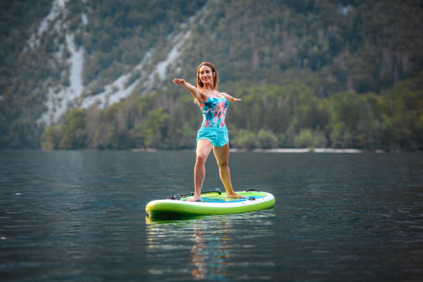 Full length front view of contented mid adult woman standing on paddleboard in Lake Bohinj doing Virabhadrasana II yoga pose.