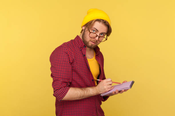 istock Hipster nerd guy in beanie hat and glasses looking at camera curious thoughtful, writing creative idea into notebook 1269869781