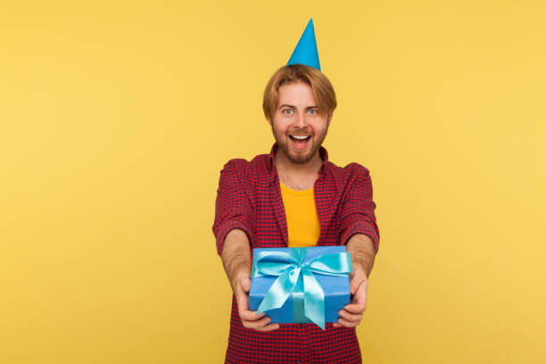 istock Cheerful delighted guy in funny party cone giving gift box, looking at camera with toothy smile, greeting 1269869802