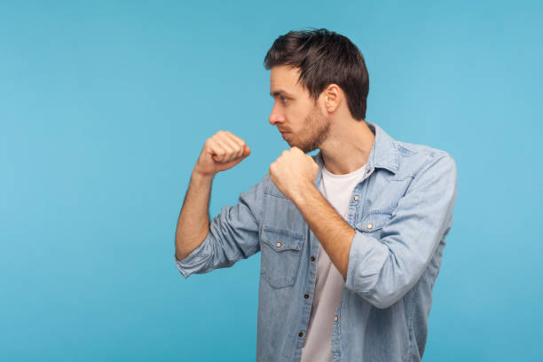 Side view of man in worker denim shirt standing with boxing gesture, punching to camera, provoking fight Side view of man in worker denim shirt standing with boxing gesture, punching to camera, provoking fight or ready to self-defense, struggle concept. indoor studio shot isolated on blue background militant groups photos stock pictures, royalty-free photos & images