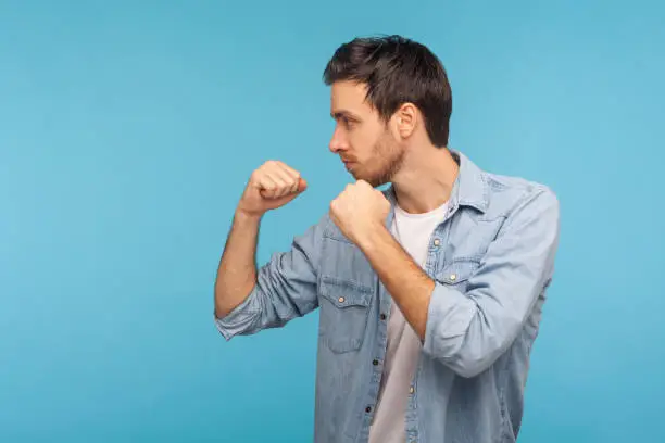 Side view of man in worker denim shirt standing with boxing gesture, punching to camera, provoking fight or ready to self-defense, struggle concept. indoor studio shot isolated on blue background